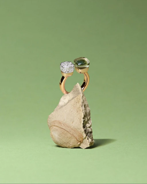 Gold ring with green and white gemstones displayed upright on a textured stone against a light green background, jewelry product shot.