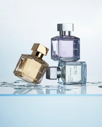 Three glass perfume bottles arranged on a reflective glass surface with water droplets against a pale blue gradient background