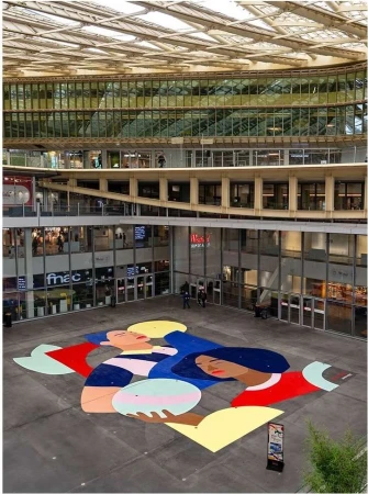 Aerial view of a large mural on a concrete surface in a covered urban space, with a building featuring layered windowed levels in the background.