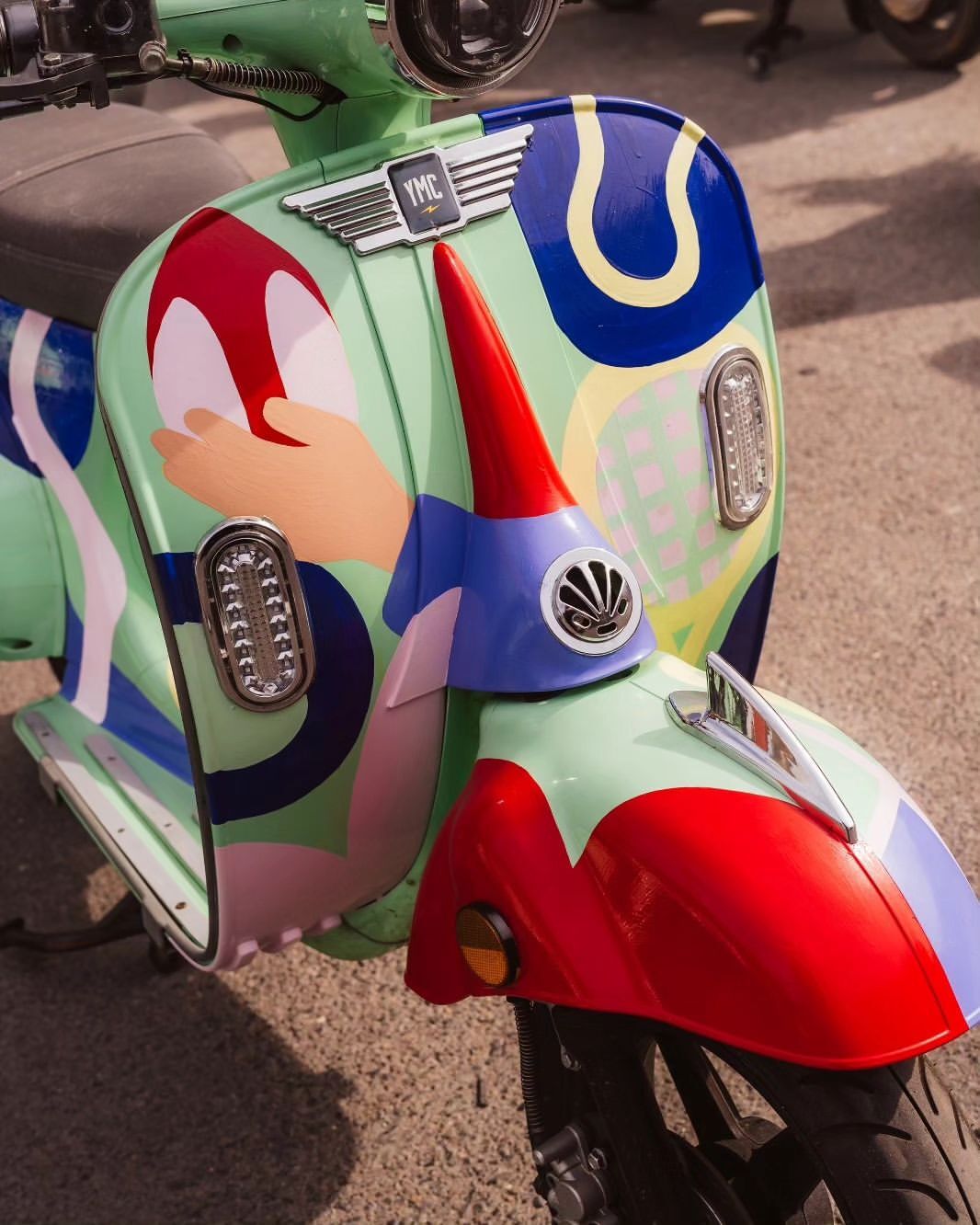 Close-up of a light green motor scooter with colorful pop-art style accents, silver trim, and a red and blue front fender.