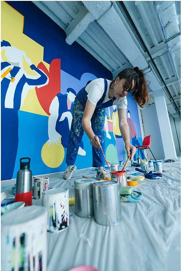 Woman in blue overalls painting a colorful mural in a spacious studio with paint cans, capturing an artistic moment in bright, natural light.