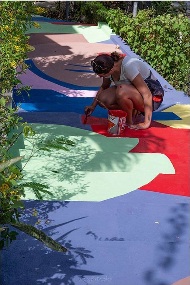 Woman artist painting colorful stairs outdoors on a sunny day. She is using a brush and a can of paint, surrounded by green plants.