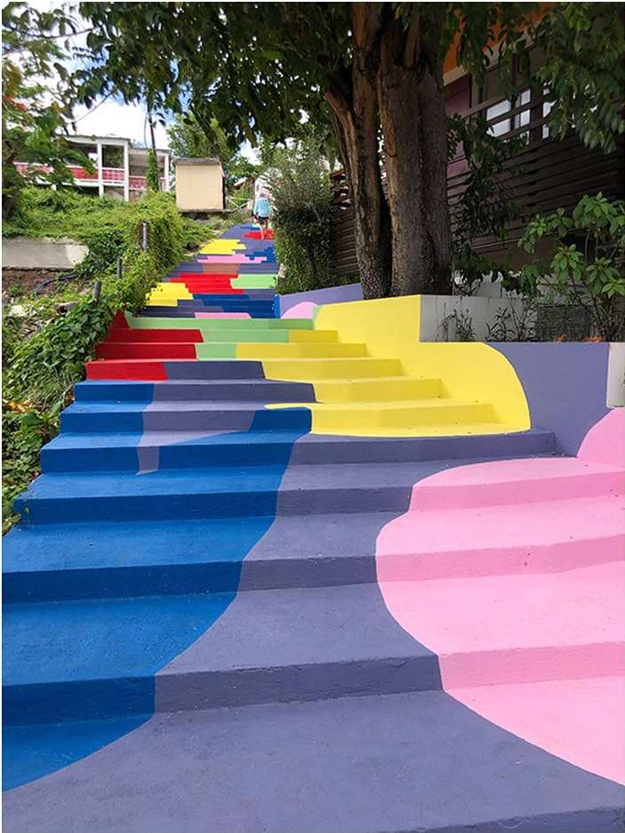 Colorful painted stairs lead upwards in an outdoor setting with a person at the top and trees lining the side of the staircase.