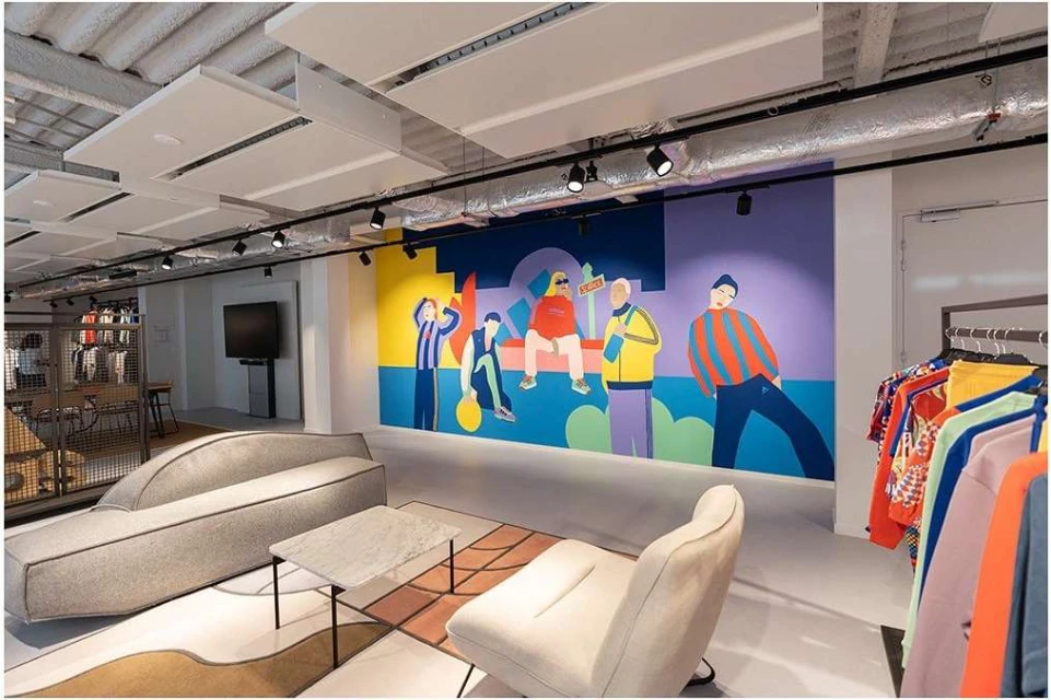 Indoor clothing store lounge area featuring a grey sofa, chair, coffee table, clothing rack, and wall art with colorful figures.