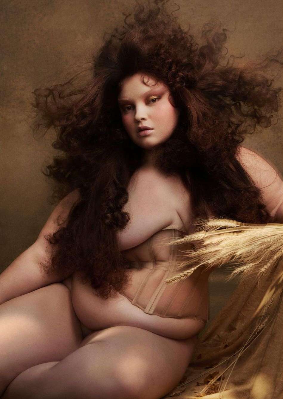 Portrait of a plus-size woman with curly brown hair and light makeup, seated and holding wheat stalks against a brown background.