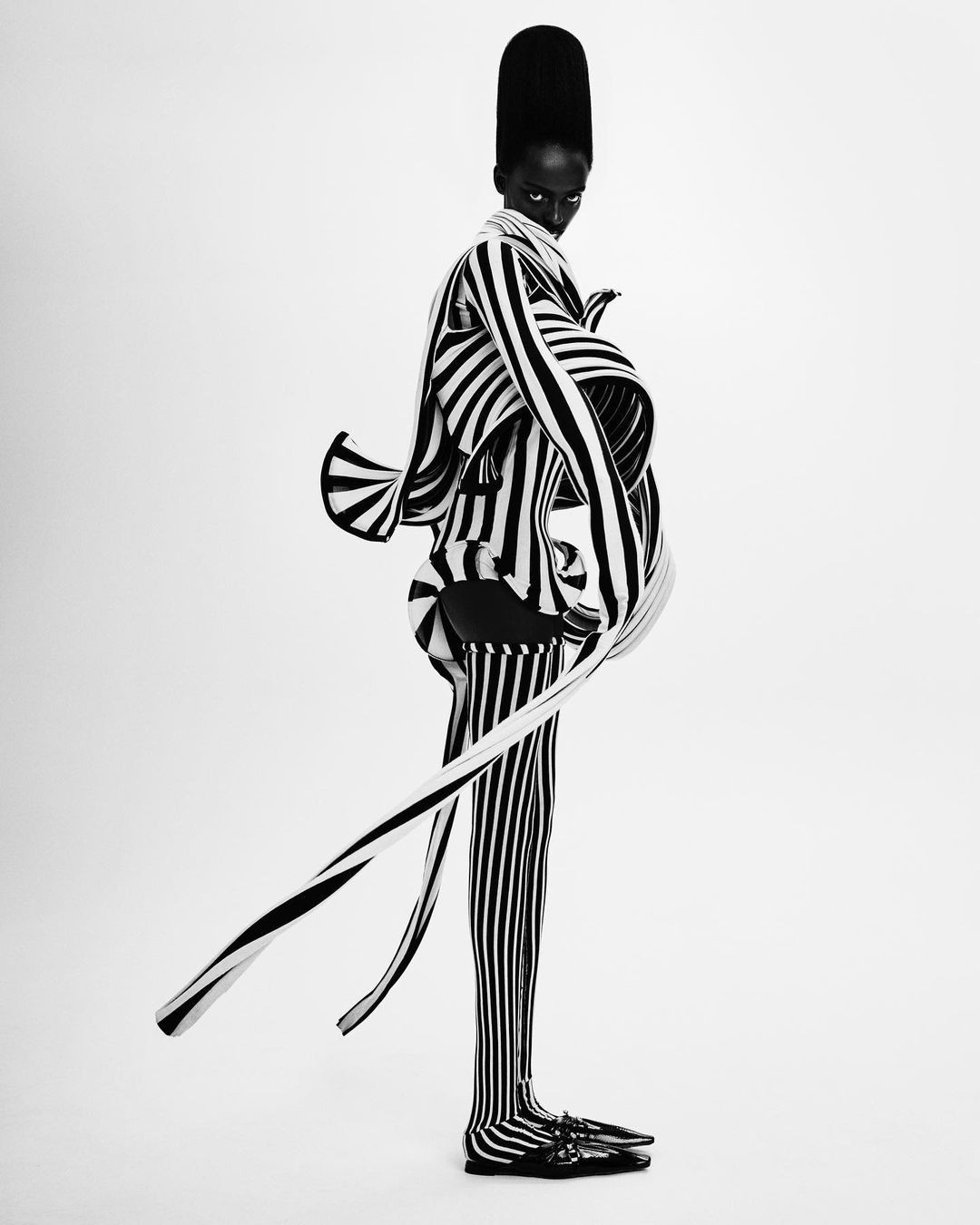 Monochrome full-body shot of a dark-skinned person in a high-fashion striped outfit against a white background in a studio setting.
