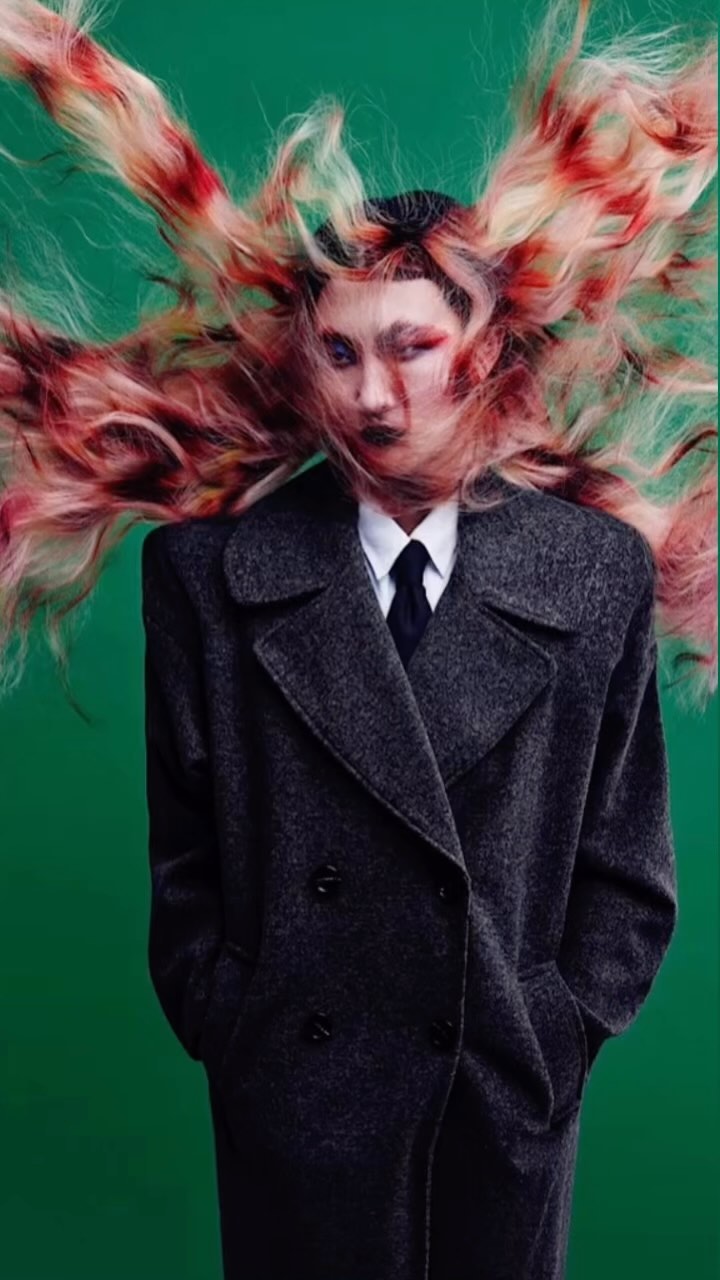 Non-binary person with red eye makeup and a gray wool coat, standing against a green background, hair styled as if caught mid-motion.