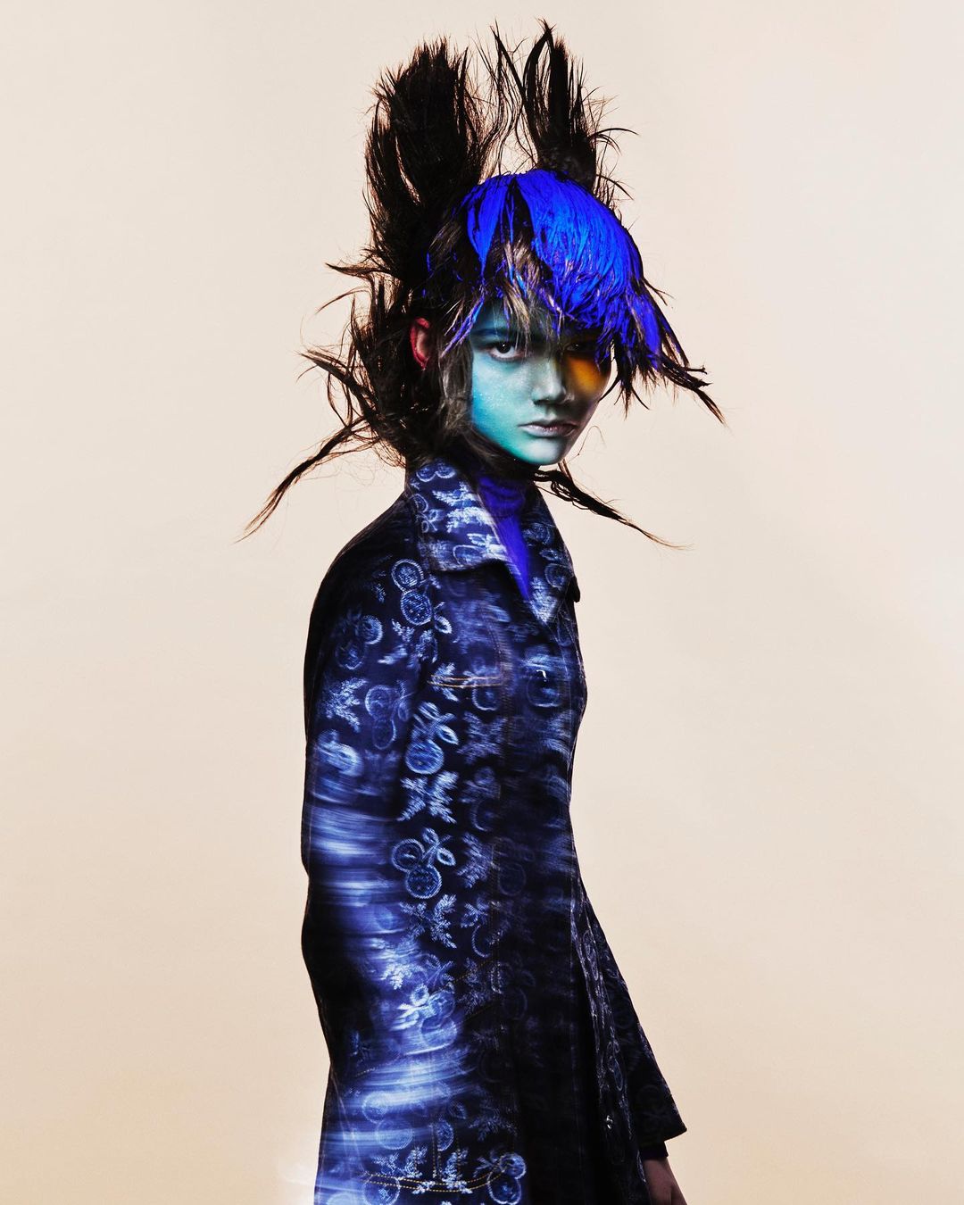 A single young adult with a blue hairstyle and painted face, wearing a patterned coat against a light background in a fashion style portrait.