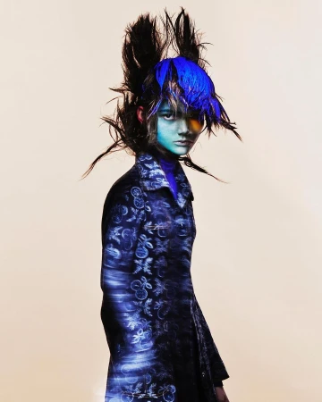 A single young adult with a blue hairstyle and painted face, wearing a patterned coat against a light background in a fashion style portrait.