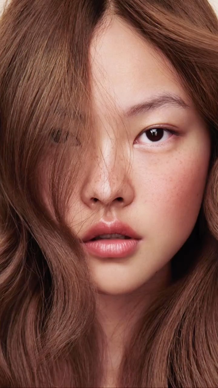 Close-up portrait of an Asian woman with medium-length wavy brown hair partially covering her face, looking directly at the camera.