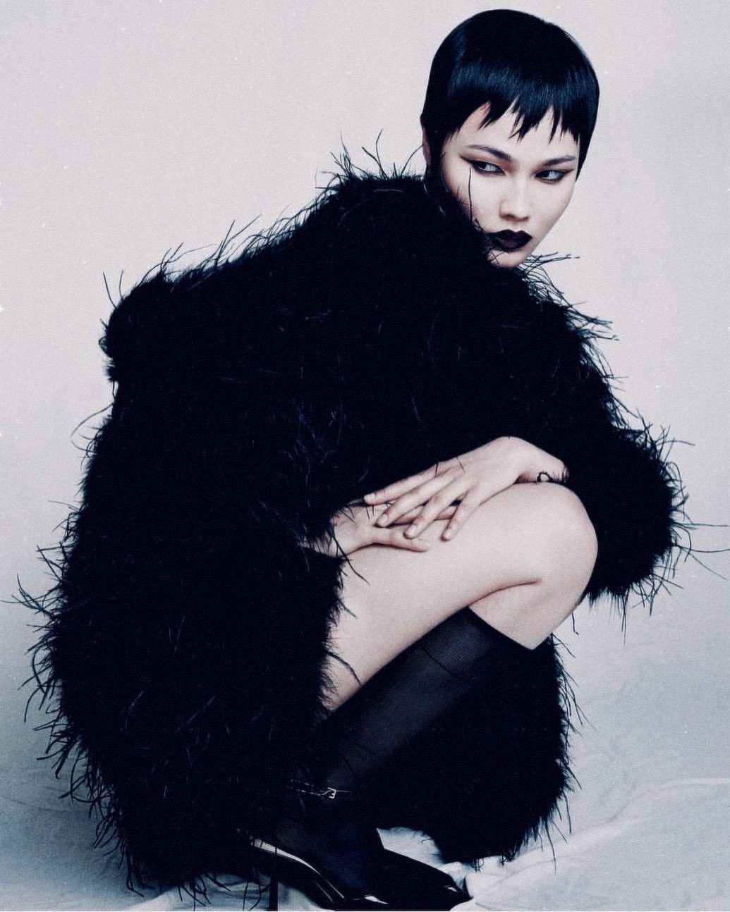 A woman with short black hair and dark lipstick poses crouched wearing a fluffy black coat, black hose, and high heels in a studio setting.