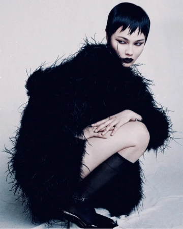 A woman with short black hair and dark lipstick poses crouched wearing a fluffy black coat, black hose, and high heels in a studio setting.