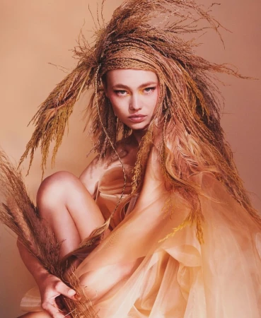 Close-up of a model with braided wheat-like hair and a sheer peach-colored dress, set against a soft peach background.