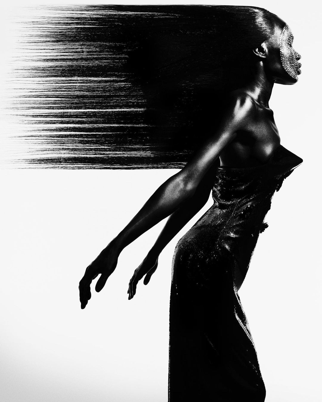 Black and white portrait of a woman in profile wearing a dress, with her long hair flowing behind her, against a white backdrop.