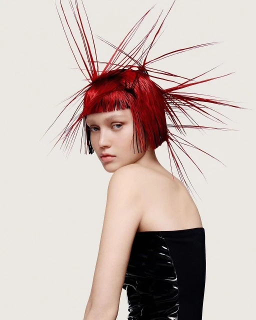 Glamour shot of a woman with vibrant red hair styled in an elaborate, spiky design, wearing a black strapless top against a white backdrop.