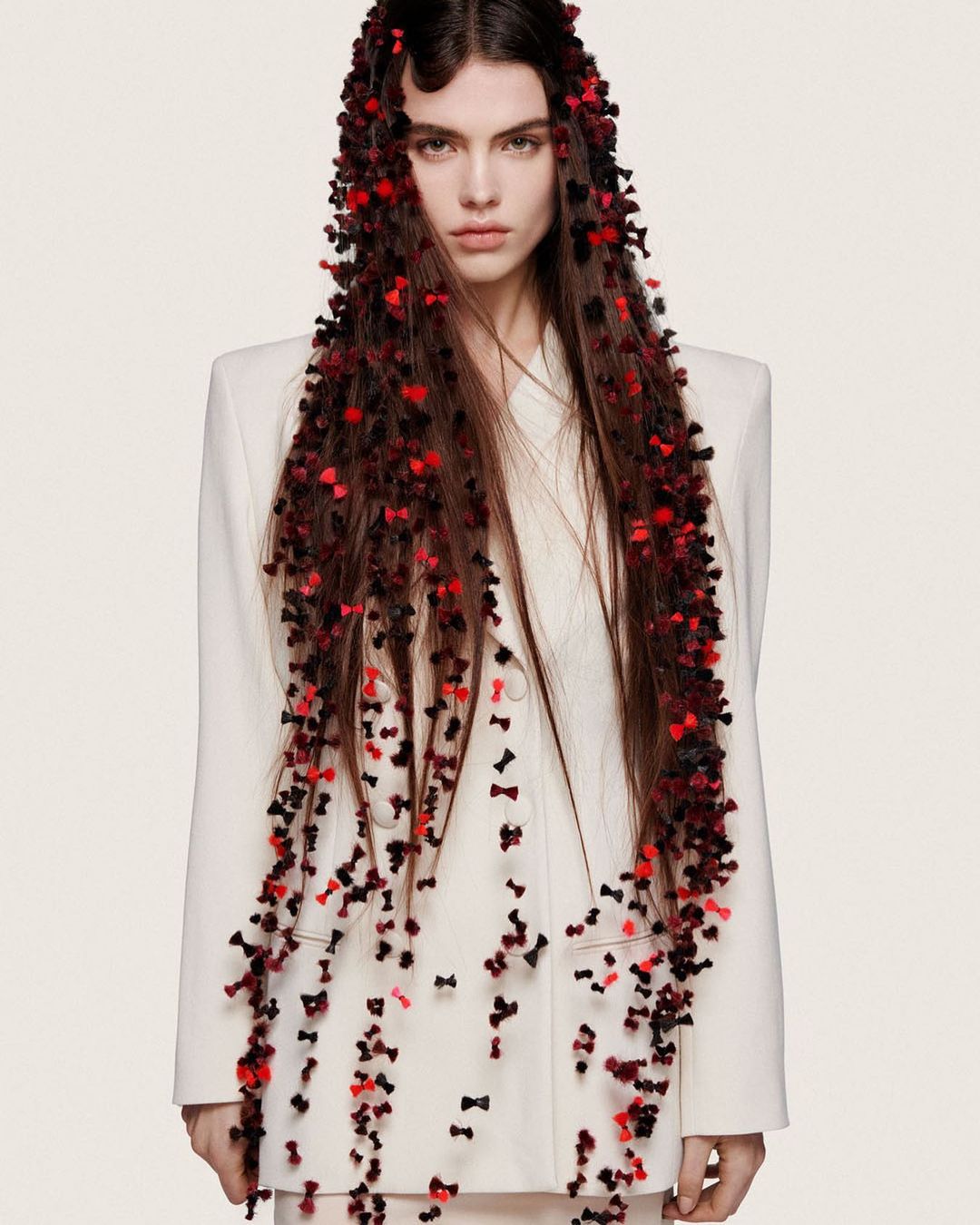 A woman with long brown hair adorned with many small black and red bows posing in front of a white background, wearing a light-colored suit.