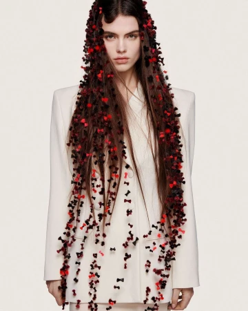 A woman with long brown hair adorned with many small black and red bows posing in front of a white background, wearing a light-colored suit.