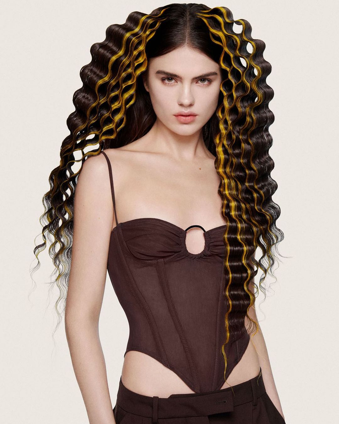 Portrait of a brunette woman with long, wavy black hair highlighted with gold, wearing a brown corset top, on a light background.