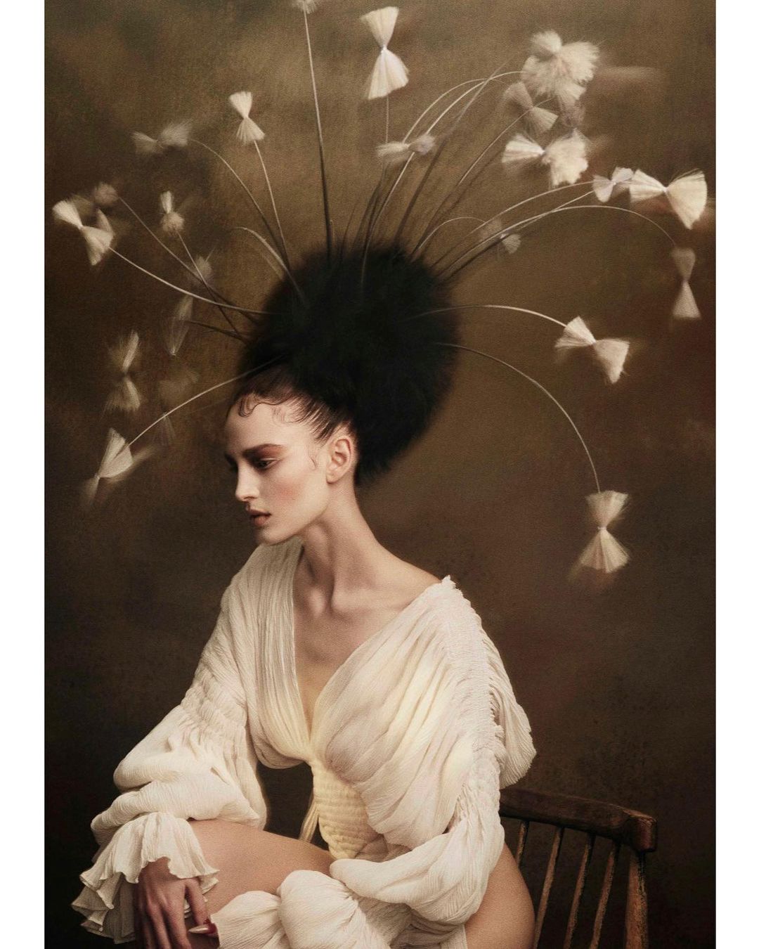 A seated woman wearing an elaborate feather headpiece and a cream dress against a dark textured background by Andrew O'Toole