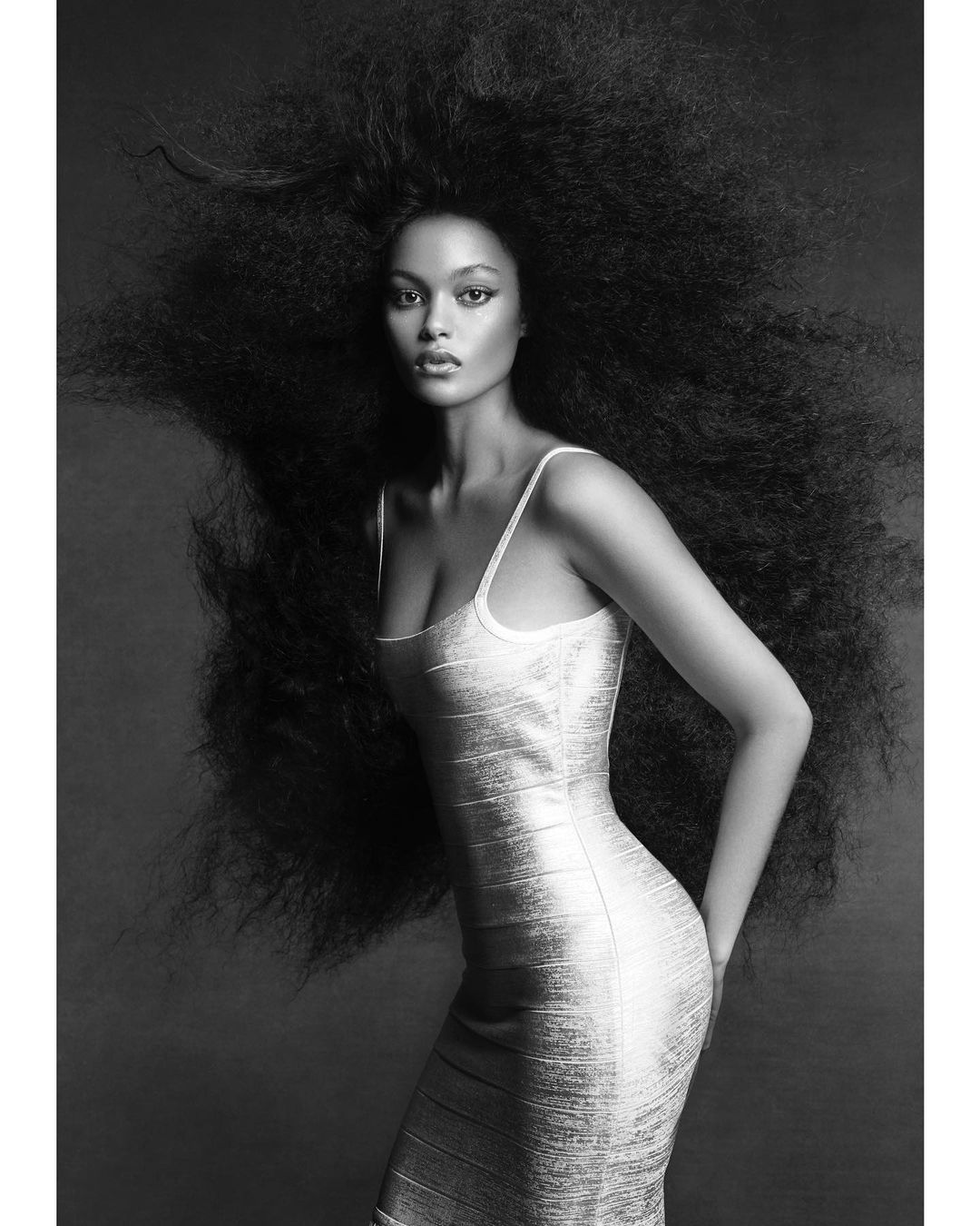 Black and white fashion portrait of a woman with voluminous Afro hair, wearing a tight dress against a dark backdrop in a studio setting.