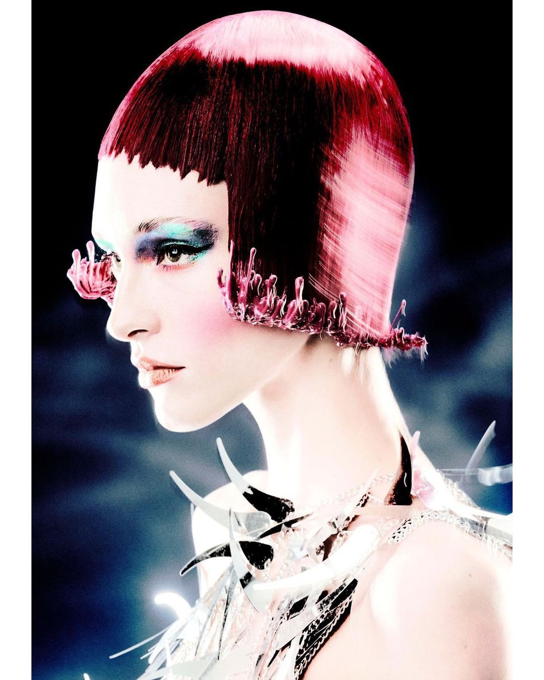 Profile shot of a woman with stylized red and pink hair, exaggerated makeup, and a decorative collar, against a blurred, dark background.