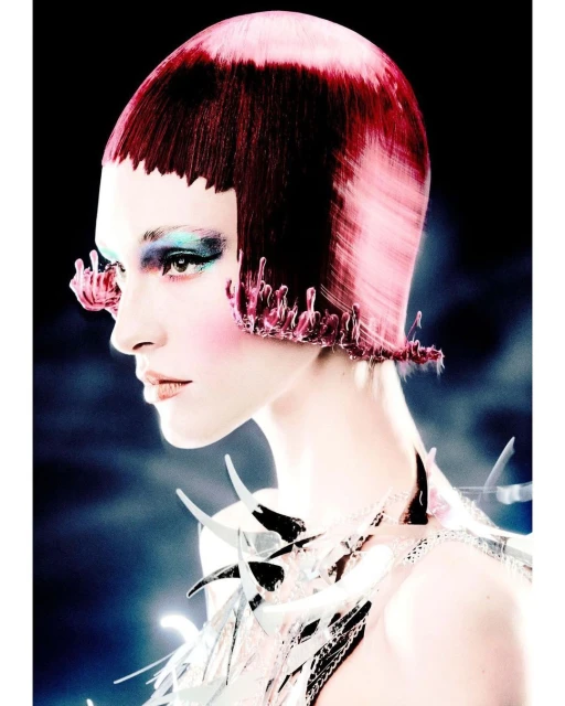 Profile shot of a woman with stylized red and pink hair, exaggerated makeup, and a decorative collar, against a blurred, dark background.
