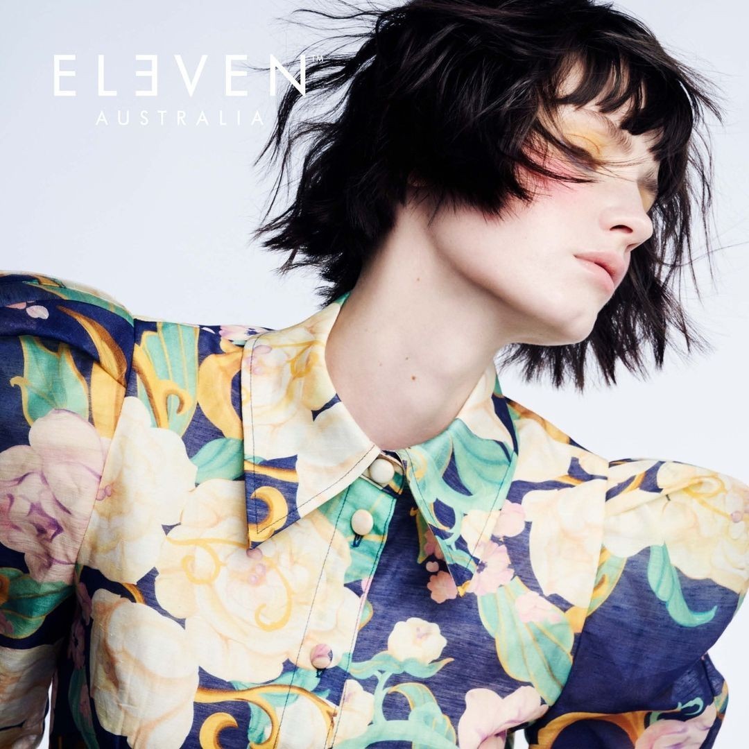 Close-up of woman with short dark hair, wearing a floral patterned blouse, eyes closed with pink eyeshadow, against a white backdrop.