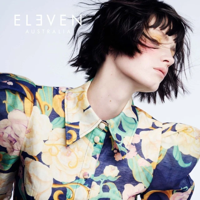 Close-up of woman with short dark hair, wearing a floral patterned blouse, eyes closed with pink eyeshadow, against a white backdrop.
