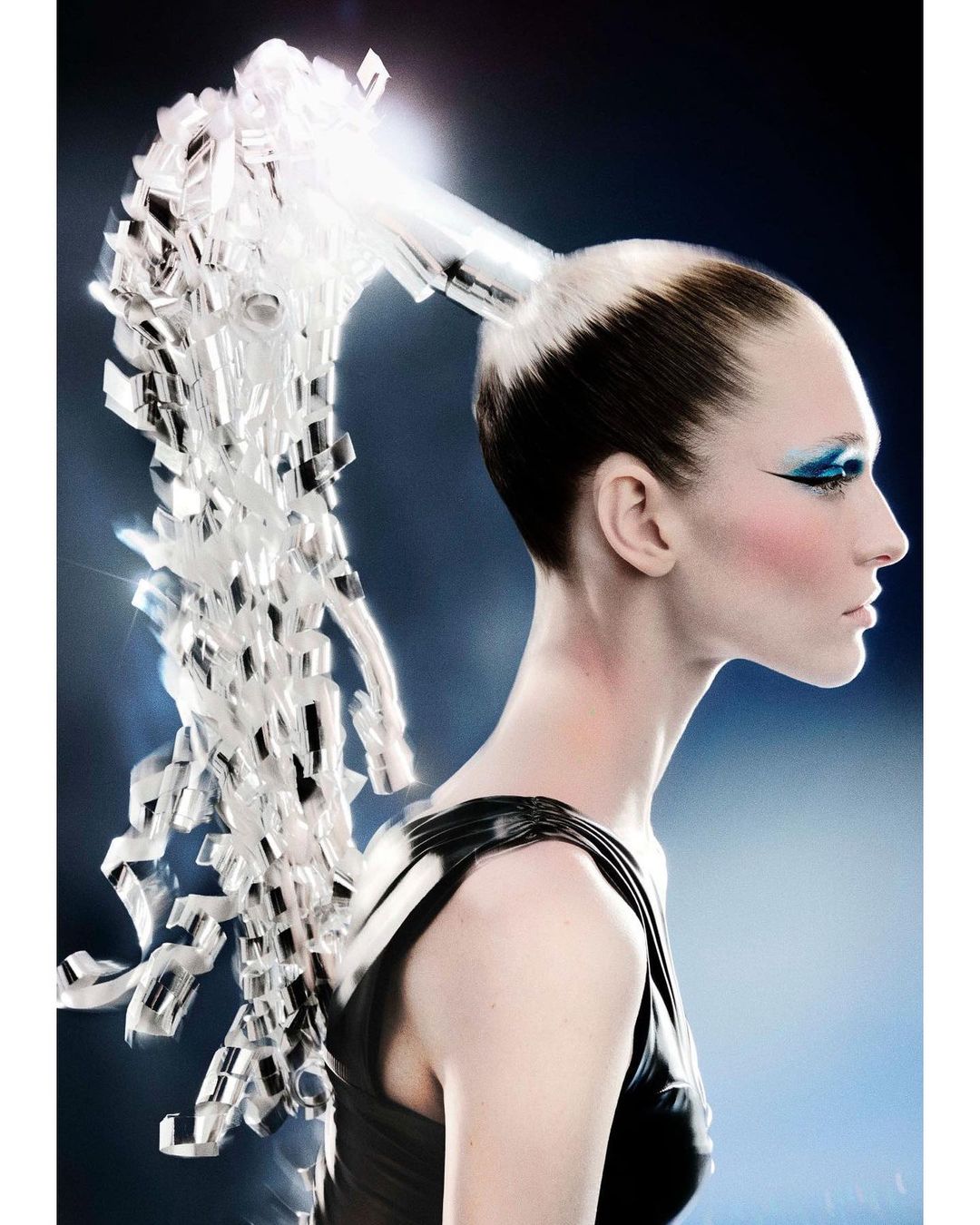 Woman with creative hairstyle and blue eyeshadow posing in a studio with a dark blue background and light shining on her hair.