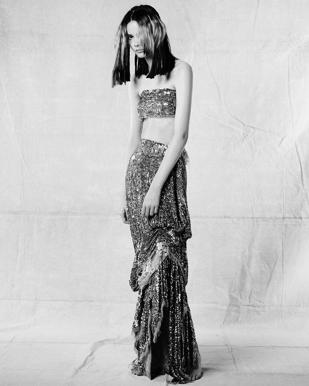 A slender fashion model with dyed hair poses in a matching shimmering sequined top and skirt in a studio setting against a crumpled backdrop.
