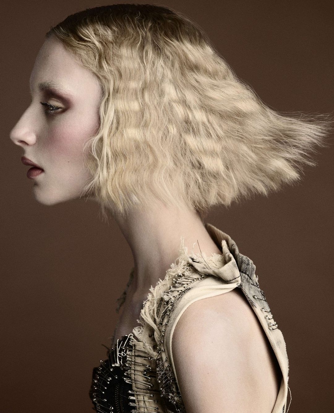 Profile shot of a woman with blonde, crimped hair, light skin, and distinctive makeup wearing a high-fashion beige dress with safety pin details.