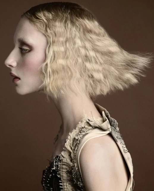 Profile shot of a woman with blonde, crimped hair, light skin, and distinctive makeup wearing a high-fashion beige dress with safety pin details.