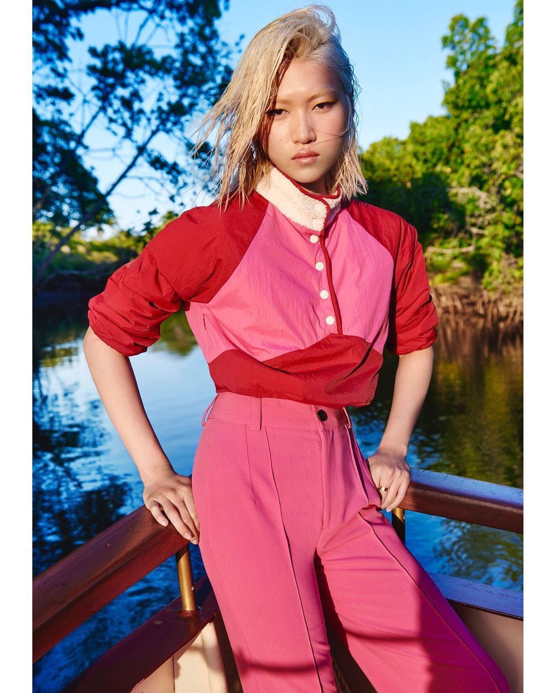 Asian woman poses in pink trousers and a color-blocked top with a buttoned collar, standing on a boat with a river and trees background.