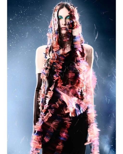 Woman in a fringed top, wearing long gloves and teal makeup, stands in a studio with a blue and black background and falling particles.