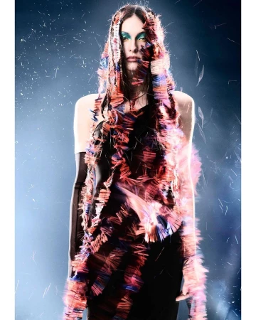Woman in a fringed top, wearing long gloves and teal makeup, stands in a studio with a blue and black background and falling particles.