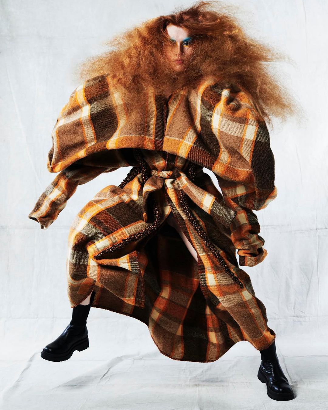 A woman with red hair in a plaid coat strikes a pose in front of a white background. The coat has puffy sleeves and is cinched at the waist.