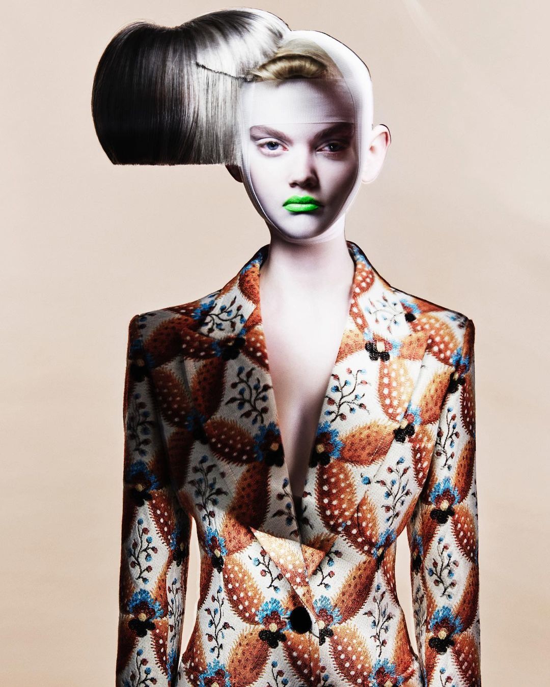 A woman with pale skin and bright green lipstick wears a patterned blazer, striking a captivating pose in a studio setting.