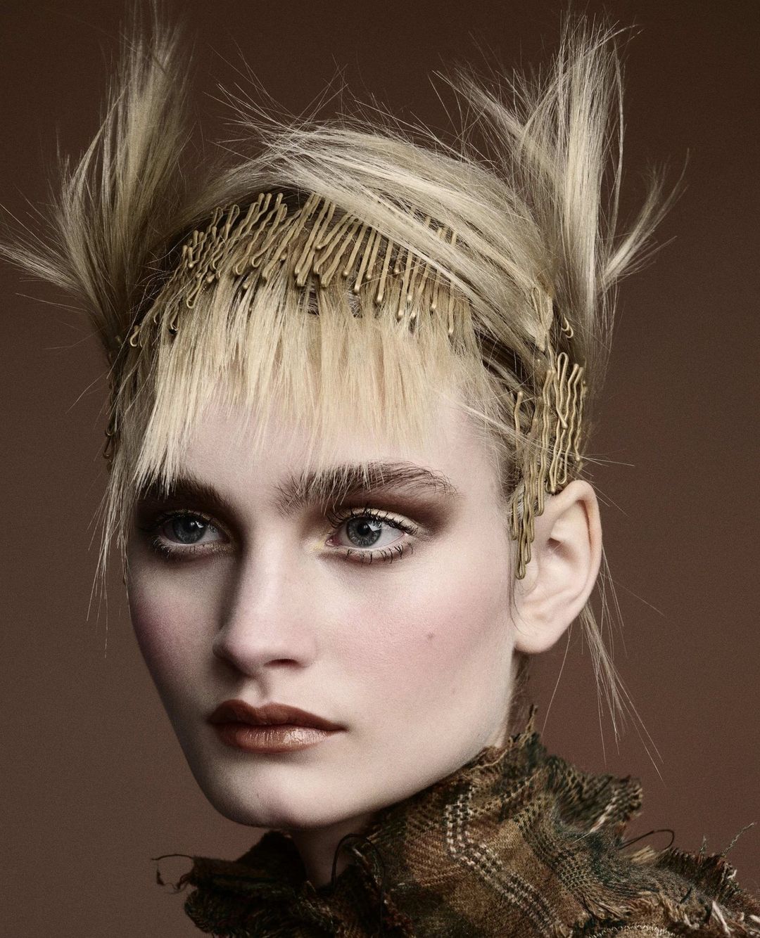 Close-up portrait of a blonde woman with creative hair styling held by bobby pins, heavy bronze makeup, and a patterned top in a studio shot.