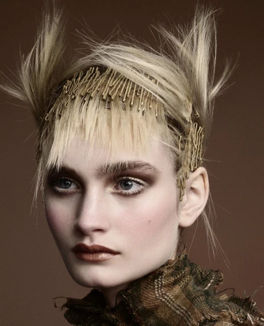 Close-up portrait of a blonde woman with creative hair styling held by bobby pins, heavy bronze makeup, and a patterned top in a studio shot.
