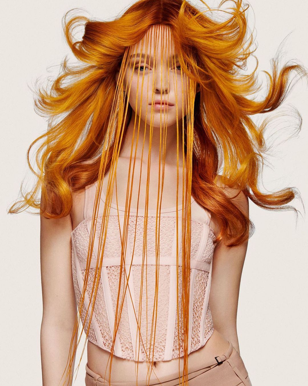 A woman with wavy, copper-colored hair posing in a light pink bustier and tan pants against a white backdrop in a studio shot.