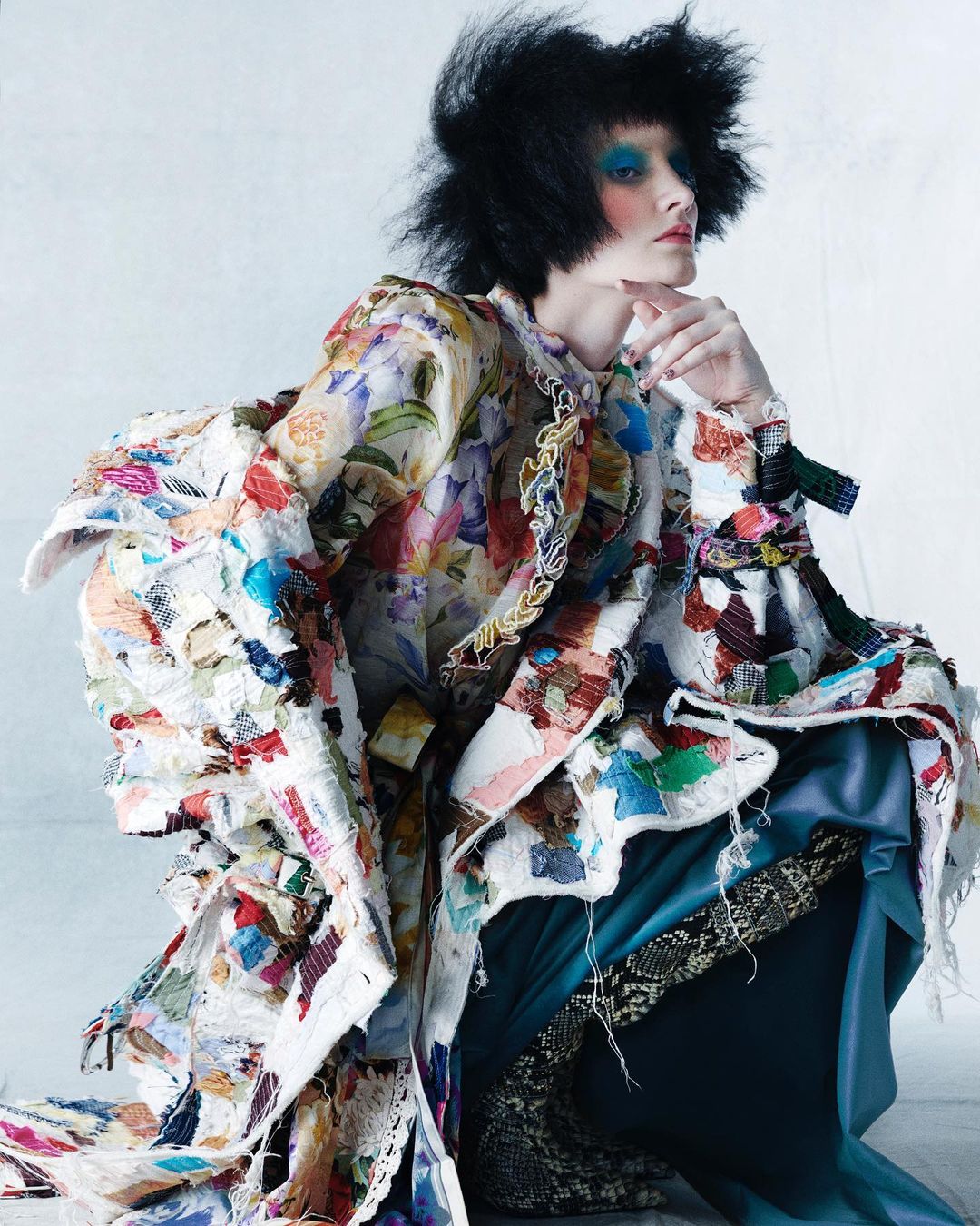 Model with dark hair poses in a studio wearing a colorful floral and patchwork jacket and teal skirt with snakeskin-print boots.