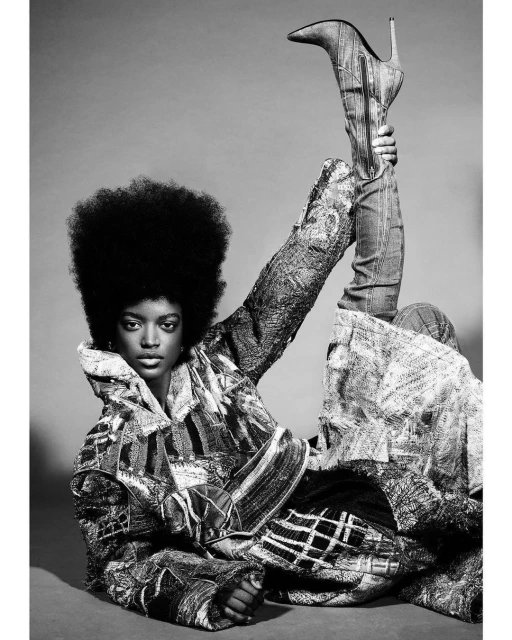 A woman with an afro poses while holding up a denim high-heeled boot in a studio shot in front of a gray background, wearing a patchwork jacket.