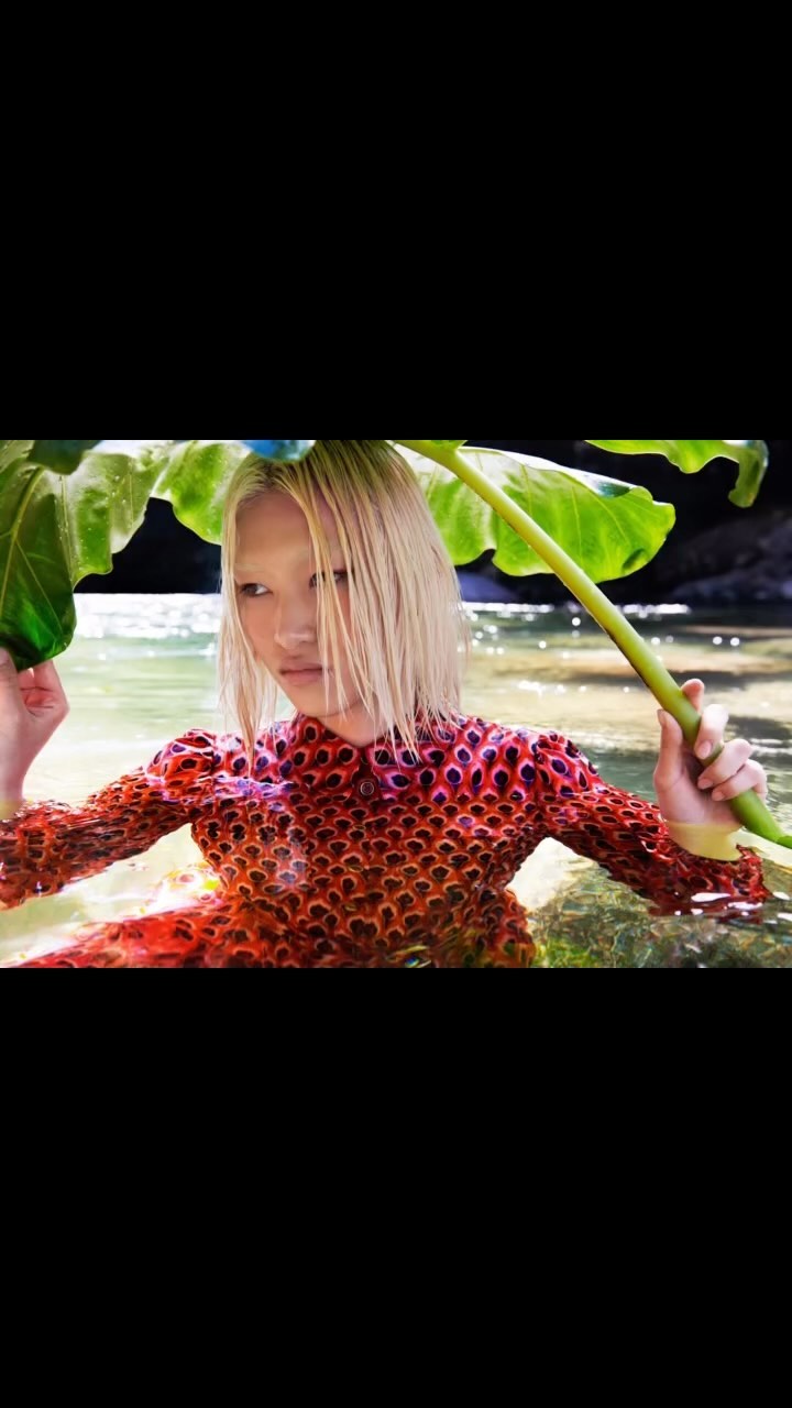 Woman in a textured dress stands waist-deep in clear water holding a large leaf over her head in an outdoor setting.