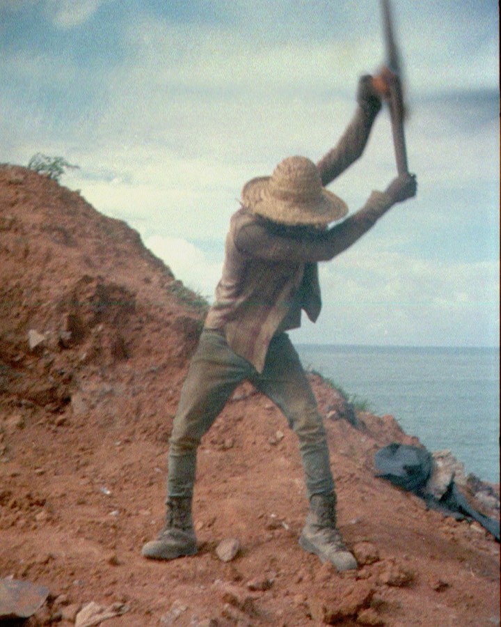 Adult male outdoors on dirt hill, wearing straw hat, light brown clothing, and boots, raising digging tool against cloudy sky and ocean.