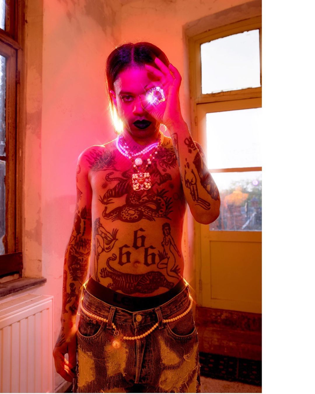 Shirtless man with tattoos, dark lipstick, and pearl jewelry poses indoors with red lighting near a window, creating a dramatic portrait.
