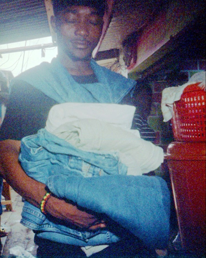 Man carrying folded clothes in indoor setting with a red plastic laundry basket visible in the background; medium shot, low lighting conditions.