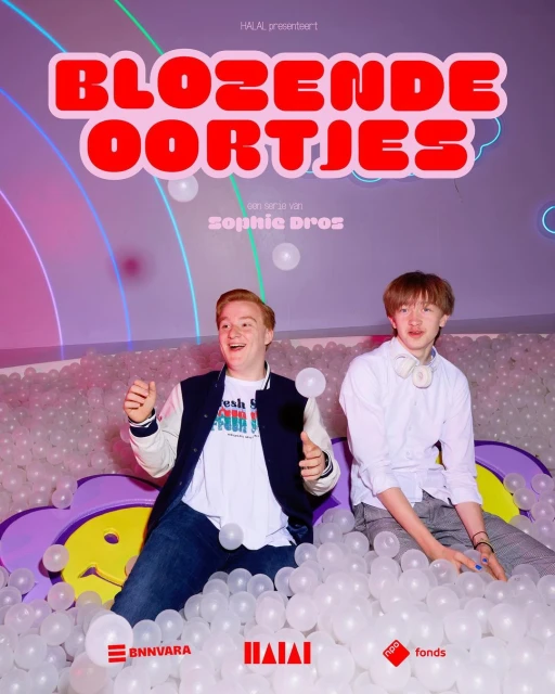 Two young men pose in a playful indoor setting filled with white balls, promoting a show called 'Blozende Oortjes' with various logos.