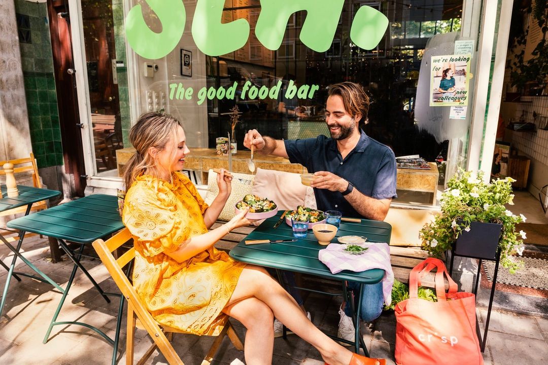 A blond woman in a yellow dress and a bearded man at a table eating salad outdoors, sharing food with a modern food bar behind them.