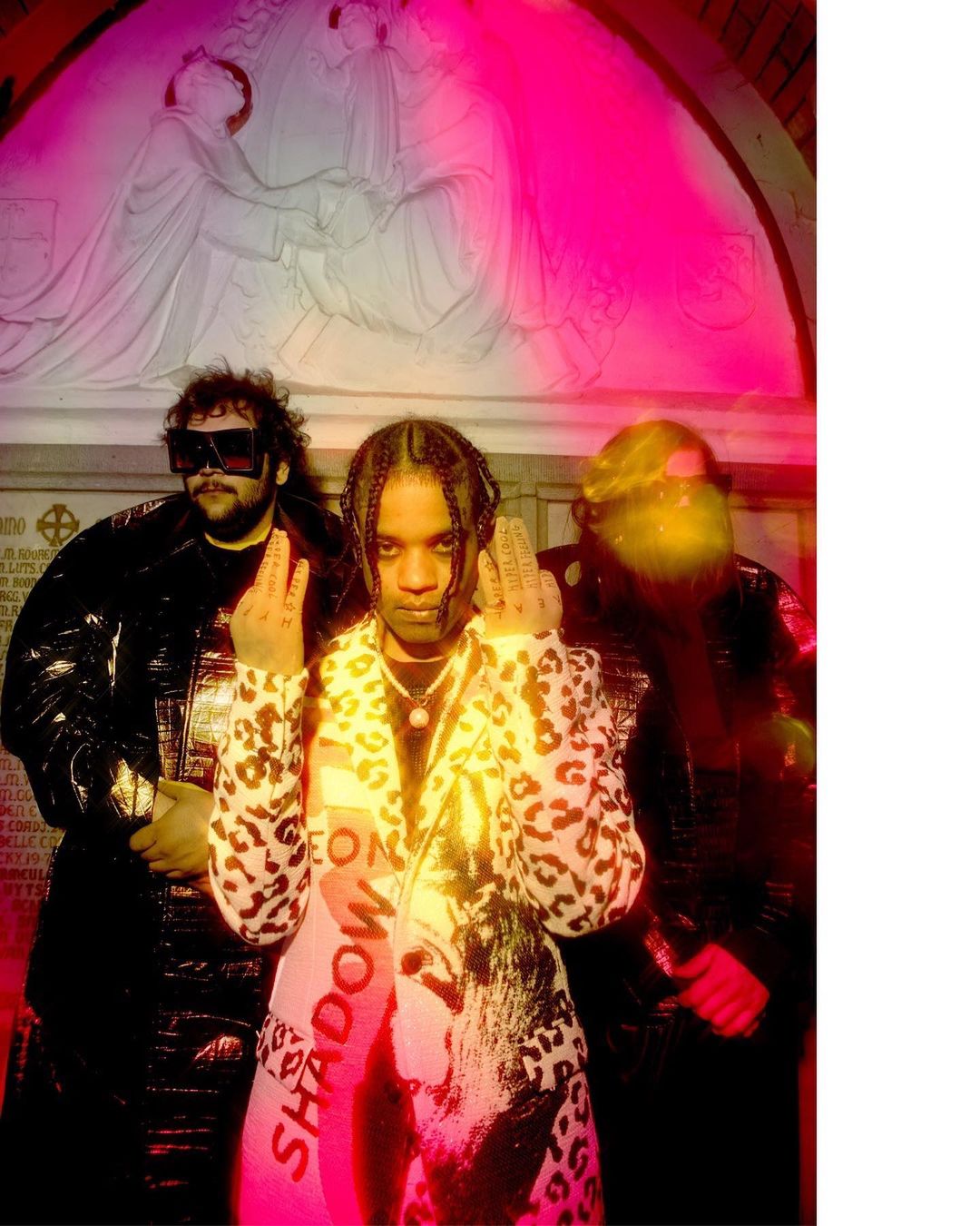 Three people pose indoors against a bas-relief wall, illuminated by pink and yellow light. One wears a leopard print coat and the others wear black jackets.