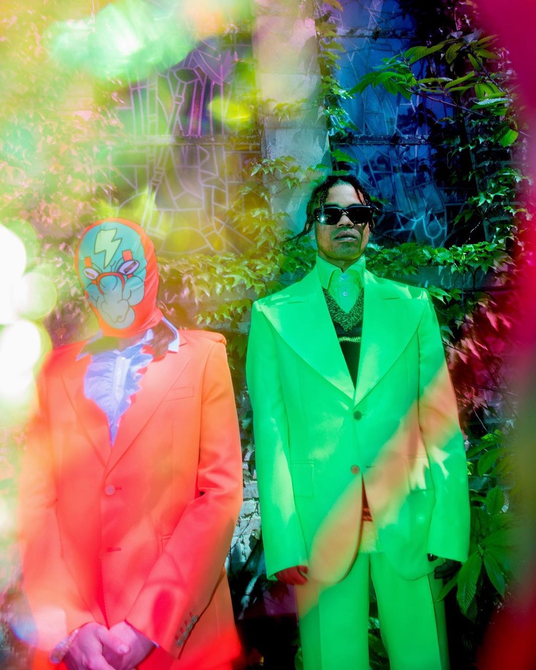 Two people stand in front of ivy-covered wall; one in neon green suit, one in orange suit with a patterned full-face mask.